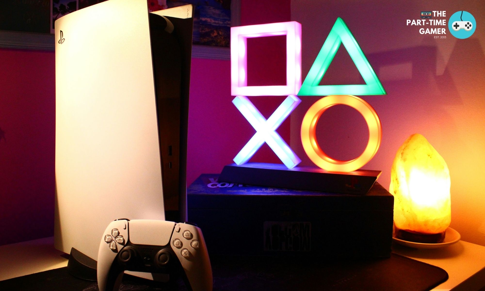 A PlayStation 5 console illuminated by warm ambient lighting next to a lamp featuring the iconic circle, square, cross, and triangle button symbols.